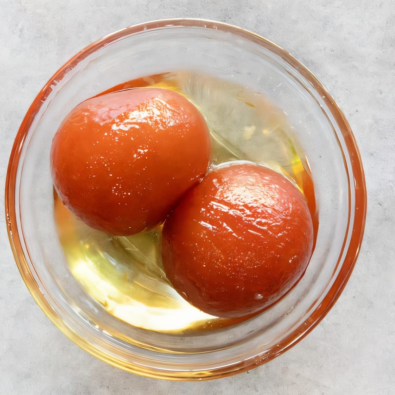 Two glistening Gulab Jamun, Indian fried dough balls, in a clear glass bowl with syrup.