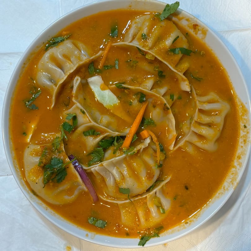 A bowl of Indian momos in a rich orange sauce, garnished with fresh cilantro and vegetables.